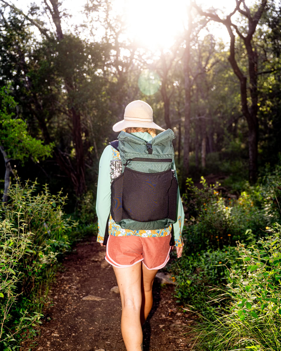 A woman walks outside with a sun hat and green backpack