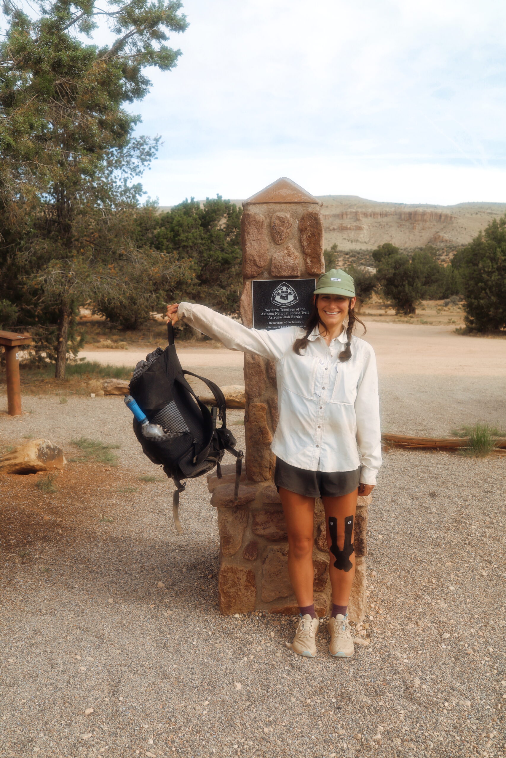 A female hiker holds up her backpack while smiling infront of the Arizona Trail terminus